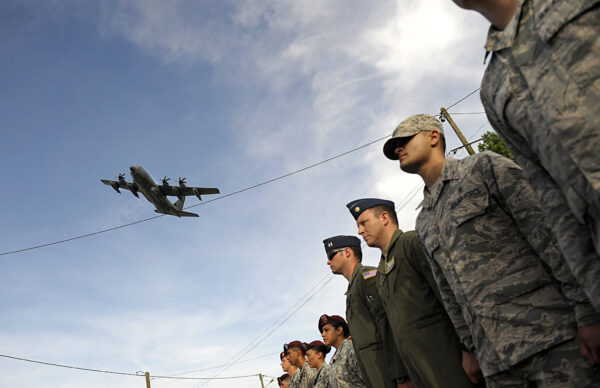 U.S. airmen standing in formation while a C-130 flies above. The ceremony honors the 70th anniversary of the D-Day invasion.